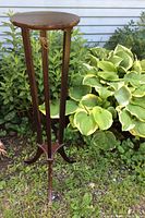 Full view of the tall wooden plant stand placed outdoors among leafy green plants, showing the 3 vertical legs and round top surface.