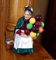 Front view of Royal Doulton figurine showing seated elderly woman holding balloons, wearing colorful clothing and flowered headscarf, with flower basket beside her.