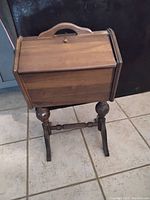 Full view of vintage wooden sewing box on wooden stand with turned legs and handle on the lid.