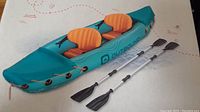 Blue inflatable kayak with orange seats and two aluminum paddles laid beside it