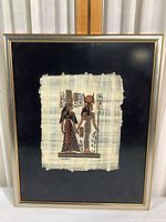 Frontal photo showing the complete framed papyrus artwork against a neutral backdrop, highlighting the frame and papyrus print centered within.