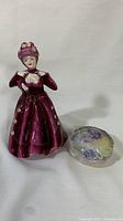 Porcelain figurine dressed in deep burgundy with small pink flowers and matching floral hat next to a floral trinket box