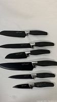 Photo of six Chef Essential knives with black coated blades and black silicone handles, arranged on a white background showing full length and handle design.