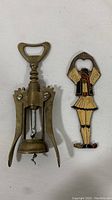 Photo showing two vintage brass items on a white cloth background: a winged corkscrew wine opener and a Danish dancer themed bottle opener.