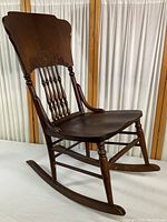 Side view of the full vintage wood rocking chair showing decorative carved backrest, turned spindle back, curved arms, and contoured wooden seat.