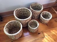 Group shot of five handcrafted woven baskets of varying sizes on wooden floor near wall.