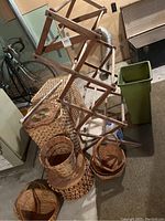 Lot of assorted woven baskets, wooden drying rack, green wastebasket, and laundry hampers in basement setting.