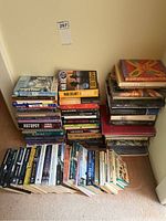 Overview of stacked books showing a variety of fiction and non-fiction titles in paperback and hardcover formats, arranged in three vertical stacks on the floor against a wall.