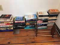 Wide view of stacks of books on a wooden floor showing multiple titles and a folded wooden bookstand leaning against the wall.