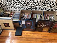 Four cardboard boxes filled with CDs and cassette tapes arranged on the floor with some audiobook tapes displayed in front.