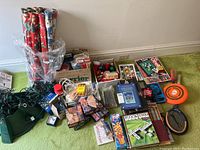Wide shot showing entire lot including Christmas wrapping paper rolls, Christmas lights, tree stand, various ornaments in boxes, and a variety of boxed and loose games such as Jenga, puzzles, Pick Two, and other family games, as well as some toy rackets and frisbee.