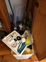 View of assorted cleaning supplies in white mop bucket with wringer on top, including Black & Decker Scrub Buster scrubber, cleaning sprays, containers and light bulbs.