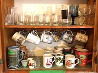 Wide shot of a cabinet shelf showing a collection of clear glass drinkware including tumblers, stemmed glasses, and martini glasses on top shelf; below are various mugs hanging and on a shelf including a tall Irish porcelain stein with scenic design