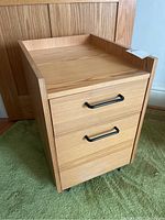 Single pressboard filing cabinet with two drawers, black handles, on casters, light wood grain finish.