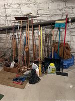 Wide shot of assorted garden tools including long-handled spades, shovels, saw, rakes, brooms, and gardening supplies against a stone wall.