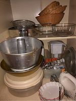Multiple woven bread baskets stacked along with glass pie plates and various cookie cutters, plastic storage containers, and a white plastic water filter pitcher. White electric hand mixer and some utensils are also visible.