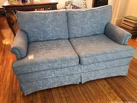 Full front view of light blue floral patterned loveseat showing two seat and two back cushions, rolled arms, and a pleated skirt base.