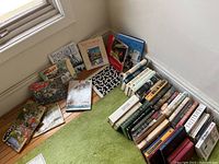 View of assorted books arranged on floor near a window, showing a variety of sizes and covers including a black and white patterned book and colorful children's or comic titles