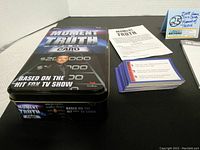 Metal game tin with 'Moment of Truth Card Game' branding and copyright from 2008 along with monetary prize graphics on lid and sides. Stack of question cards, instruction booklet, dice, and paper scorepad displayed next to tin.