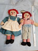 Raggedy Anne and Raggedy Andy cloth dolls laying side by side next to a measuring tape for scale, about 19 inches tall each