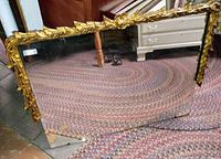 Full view of rectangular antique mirror with gold edged floral leaf carved frame top and sides resting on braided rug, showing bottom corner chip on mirror surface.