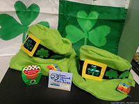 Two new green felt hats with shamrock designs and attached tags, displayed with a shamrock flag background.