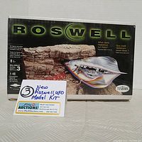Front view of sealed Testors Roswell UFO model kit box with artwork depicting the UFO crash scene and soldiers at site, includes auction info card.