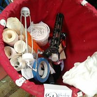 Wicker basket lined with red fabric contains wallpaper rolls, cloth rags, roller handles, painter tape, and roller cages.