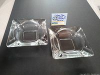 Two vintage clear glass ashtrays side by side on a white surface with auction lot sign in background.