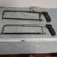 Two vintage coping saws placed side by side with measurement tape showing approximately 18 inches length. One saw has a rusty metal frame and black handle with paint wear. The other saw has a polished metal frame and black handle in better condition.