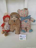 Lot of three vintage dolls: Hollie Hobbie Carrie doll in red floral dress, cloth military soldier doll, and floral cloth teddy bear with blue bows and paw pads.