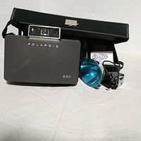 Front view of black Polaroid 330 instant camera with flash, blue flashbulb holder, and black carry case positioned behind.