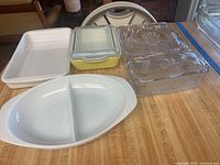Photo of all items in the lot showing white divided casserole, yellow Pyrex dish with clear ribbed lid, and two clear ribbed dishes with embossed vegetable lids on a countertop.