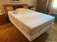 Front angle view of bed showing the white Tempur-Pedic mattress on a cream patterned box spring with headboard background and pillow on mattress.