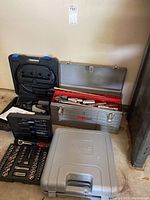 Wide view of the lot showing the Dremel rotary tool case, Craftsman metal tool chest with socket wrenches, and the closed Ryobi drill case.