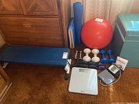 Photo showing full lot with bench, balance ball, dumbbells, ankle weights, scale, blood pressure monitor, and exercise mat on brown carpet near wooden furniture and teal storage container.