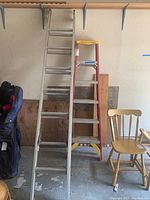 Two ladders standing side by side: red/yellow 6 ft step ladder and silver 7 ft extension ladder next to a chair and golf bag