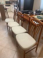 Four teak dining chairs lined up in kitchen showing front and side profiles with stained white cushions and vertical slat backs.