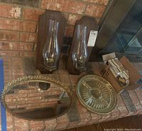 Overview showing two wood wall sconces with glass chimneys, an oval mirror tray, and a round glass tray with gold-tone rim arranged on brick surface.
