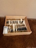 Full set of Thai ornate serviceware including spoons, forks, and knives in wooden tray, some tarnish visible.