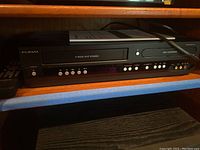 Front view of Funai DVD recorder and VCR combo unit sitting on a wooden shelf with power cord laid to the side.