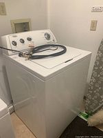 Full view of white Whirlpool top load washer with hose laid on top, showing overall cabinet and size.