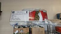Box of 7.5 ft Overland LED Pre-Lit Christmas Tree stored on metal rack, showing brand and product details on label.