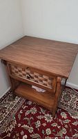 Front and angled view of wooden end table showing basket-weave textured drawer front with knob, visible open shelf area below, and wood grain finish on table top and frame.