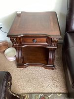 Front view of wooden Ashley Furniture end table next to dark leather seating, showing drawer and ornate carved details.