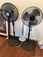Photo showing two black Lasko pedestal fans with circular bases and a white Aloha Breeze tower fan, all standing on the floor next to a wall.