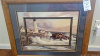 Full view of framed bilevel art print depicting lighthouse and coastal scene with white lighthouse, buildings, and crashing waves in wooden frame.
