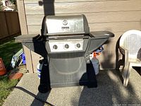 Outdoor propane BBQ grill with side burner and three control knobs, showing the full grill with side shelves and wheels.