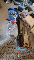 Overall view of saw, Fiskars hedge trimmers, blue bin, plastic container, and various small items on carpeted floor