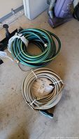 Two coiled garden hoses on the ground, one green with yellow stripes and one white in packaging, alongside a black hose holder.